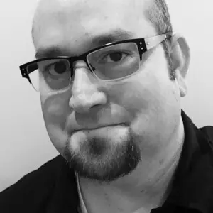 A person with short hair, glasses, and a goatee looks at the camera and smiles slightly. The black-and-white image features subtle elements inspired by Pacific Northwest custom home architecture in the background.