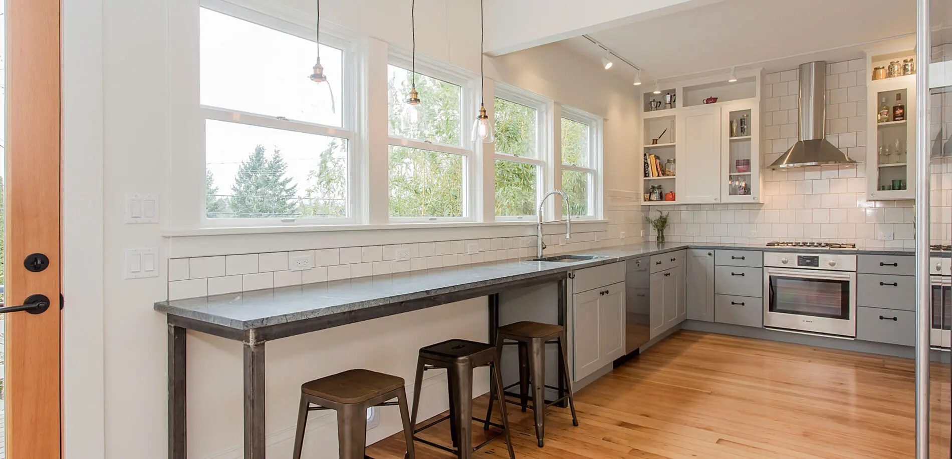 Modern kitchen with gray cabinets, stainless steel appliances, bar seating with stools, and a white subway tile backsplash—showcasing Greater Seattle custom home design—with large windows letting in natural light.