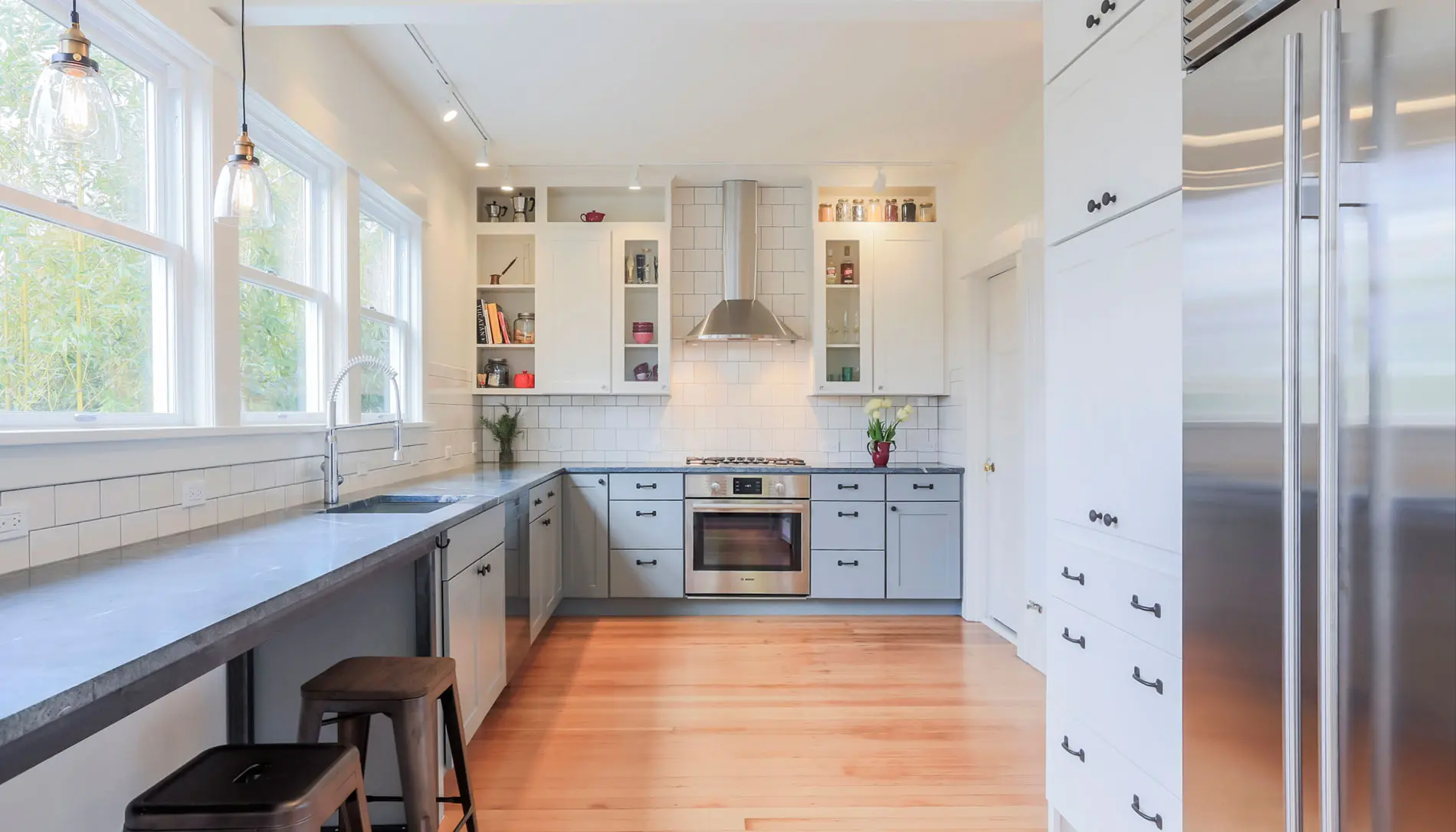 Modern kitchen with light blue cabinets, stainless steel appliances, white tile backsplash, and large windows. Light wood flooring and counter seating reflect the sleek elegance of Pacific Northwest luxury residential architecture.