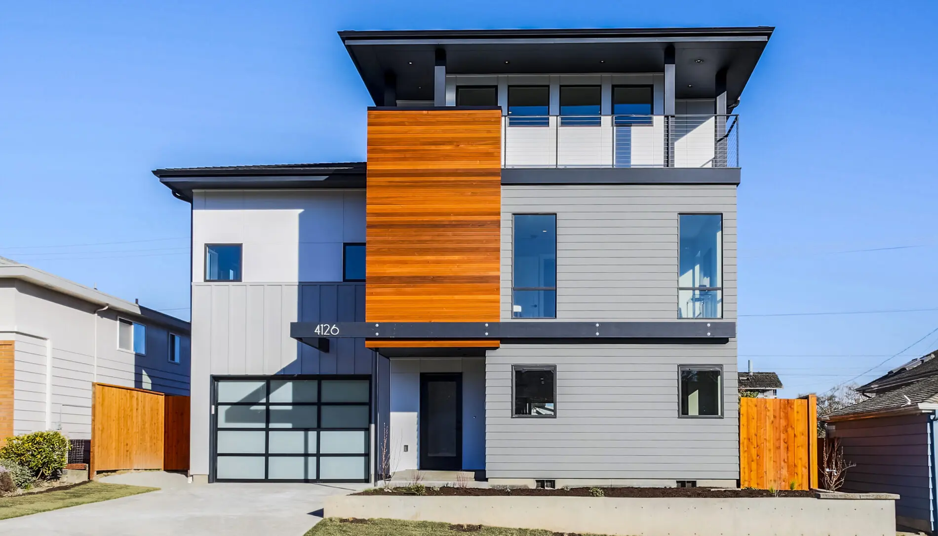 Modern two-story house with gray siding, large windows, wood panel accents, and a glass garage door showcases West Seattle custom home design on a small front lawn under a clear blue sky.