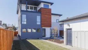 Modern three-story West Seattle custom home design featuring gray and wood paneling, large windows, and a small landscaped front yard next to a white garage.