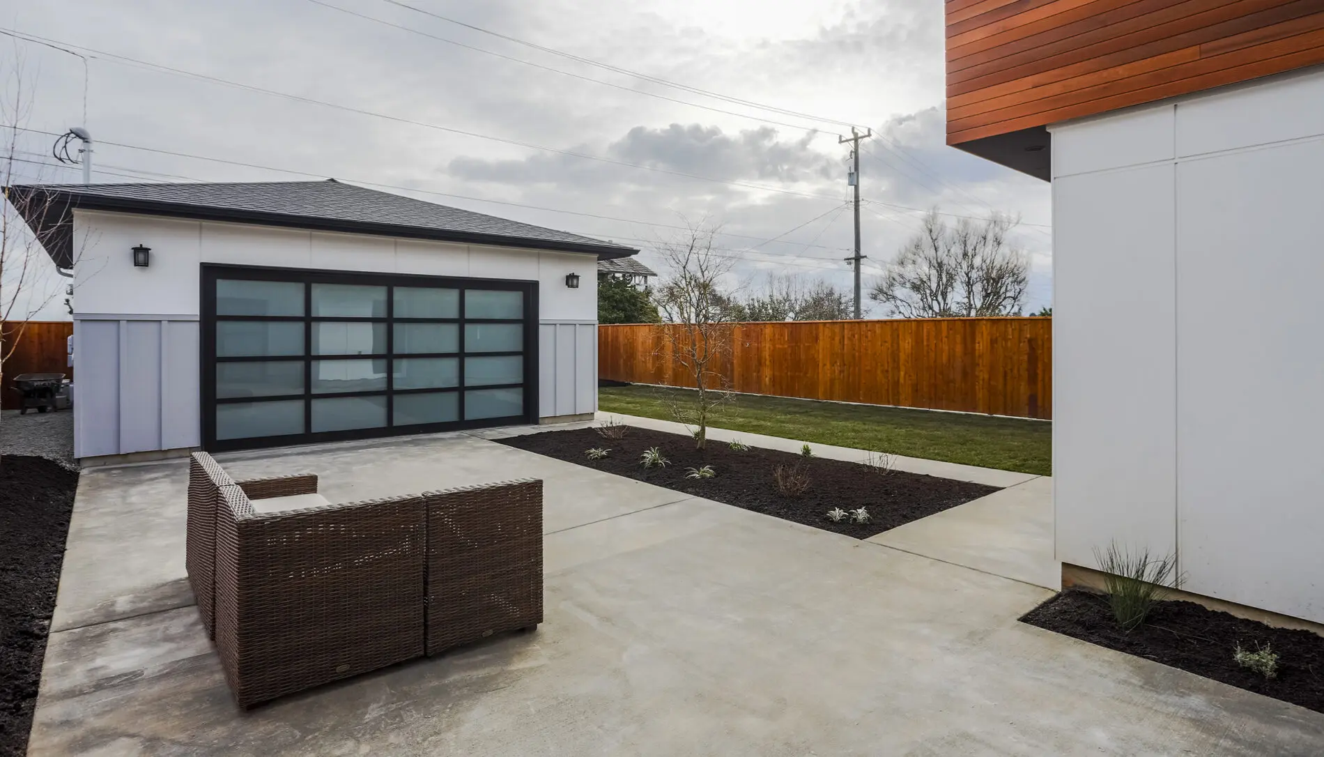 Modern backyard with a paved patio, brown outdoor seating, minimalist landscaping, detached garage with frosted glass door, and a wooden privacy fence—showcasing sleek West Seattle custom home design.