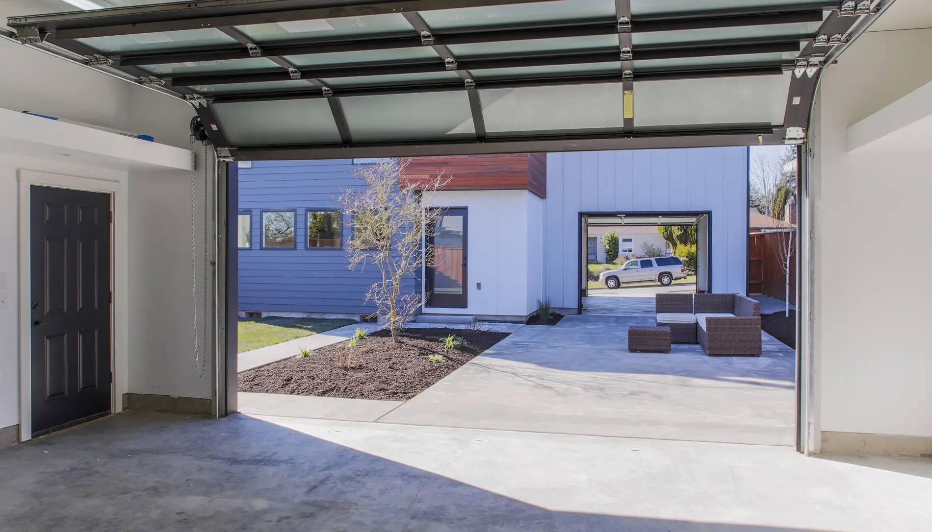 View from inside a garage with an open door, looking out onto a modern West Seattle custom home design, patio furniture, and driveway with a parked vehicle in the distance.