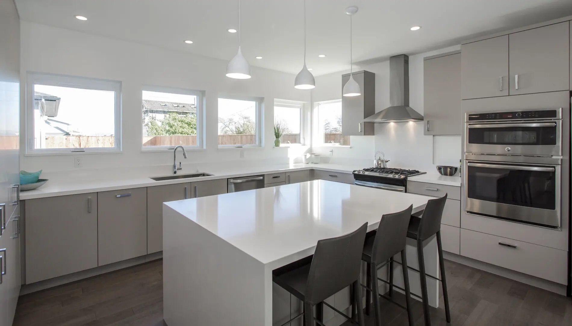 Modern kitchen with light gray cabinets, stainless steel appliances, a large white island with three dark chairs, and pendant lights; natural light fills the space in this West Seattle custom home design.