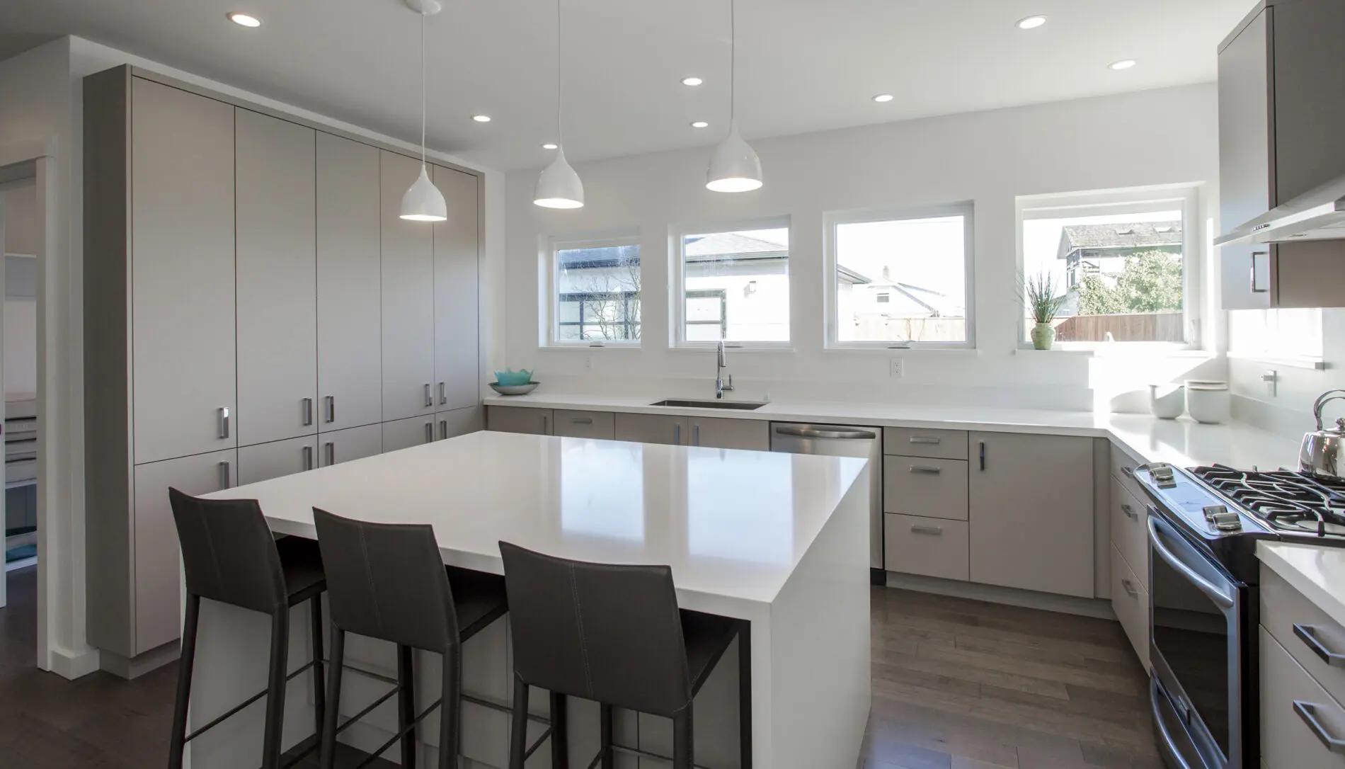 Modern kitchen with light gray cabinets, white countertops, an island with three chairs, pendant lights, and three windows letting in natural light—showcasing the elegance of West Seattle custom home design.