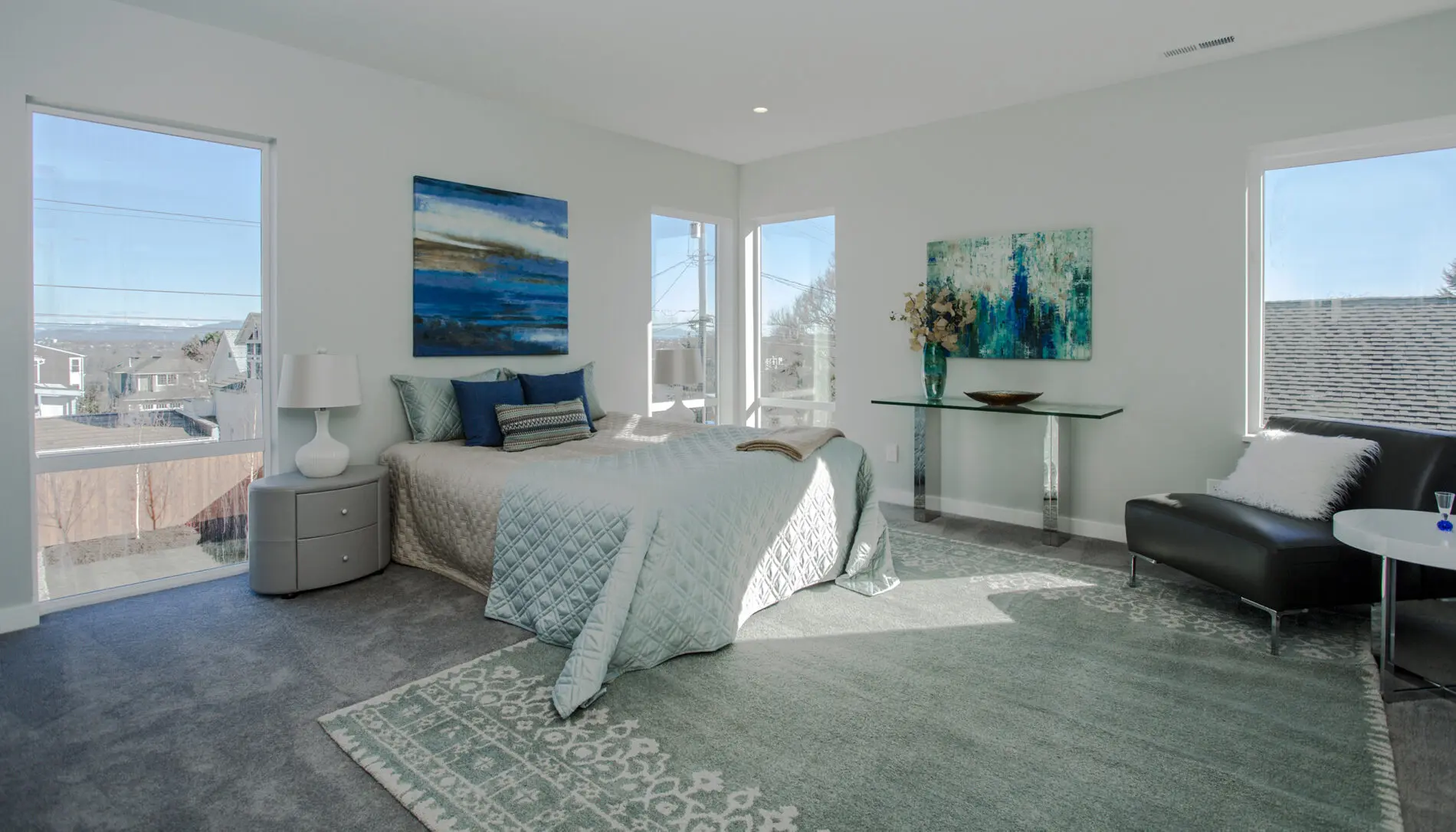 Modern bedroom in a West Seattle custom home design, featuring large windows, a bed with blue and gray bedding, contemporary artwork, a glass console table with flowers, and a gray chair on a patterned rug.