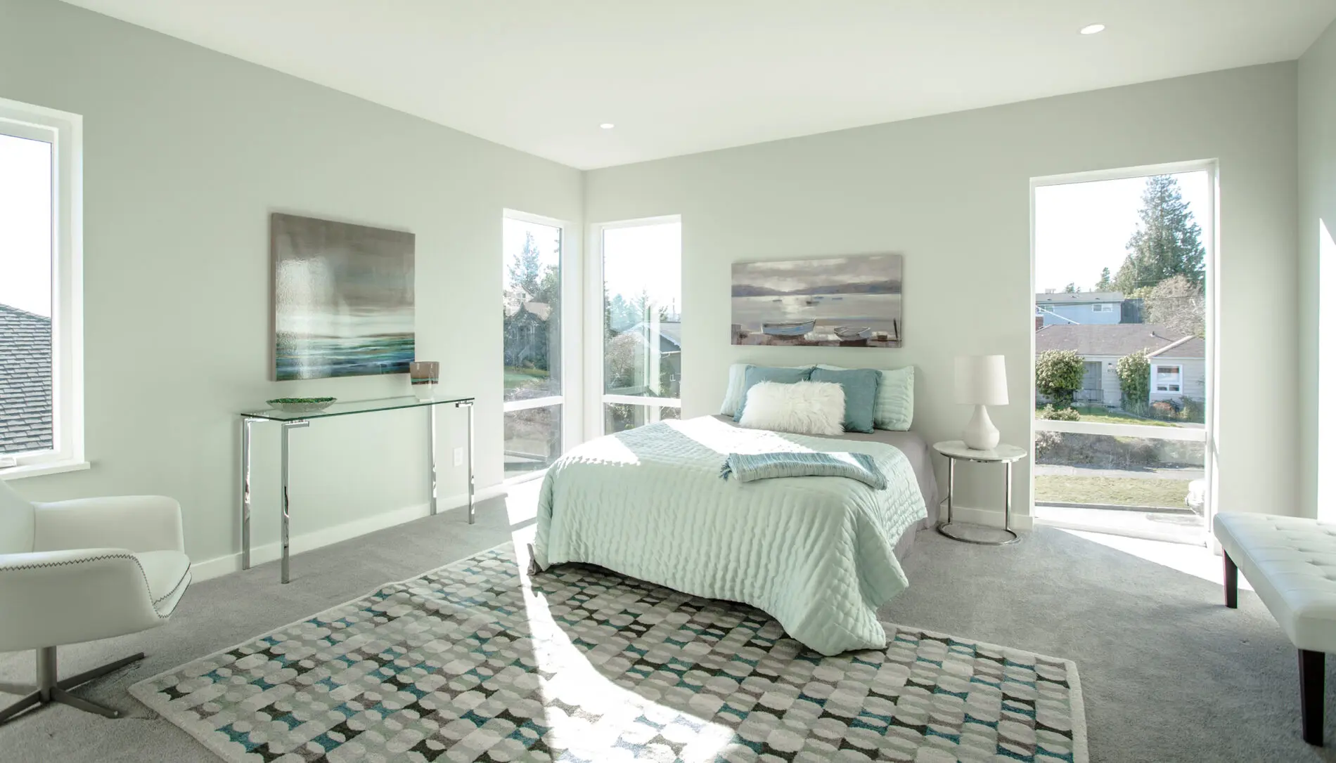 Bright bedroom with large windows, a bed with light blue bedding, a patterned rug, a chair, glass desk, and landscape paintings—showcasing the airy feel and thoughtful details of West Seattle custom home design.