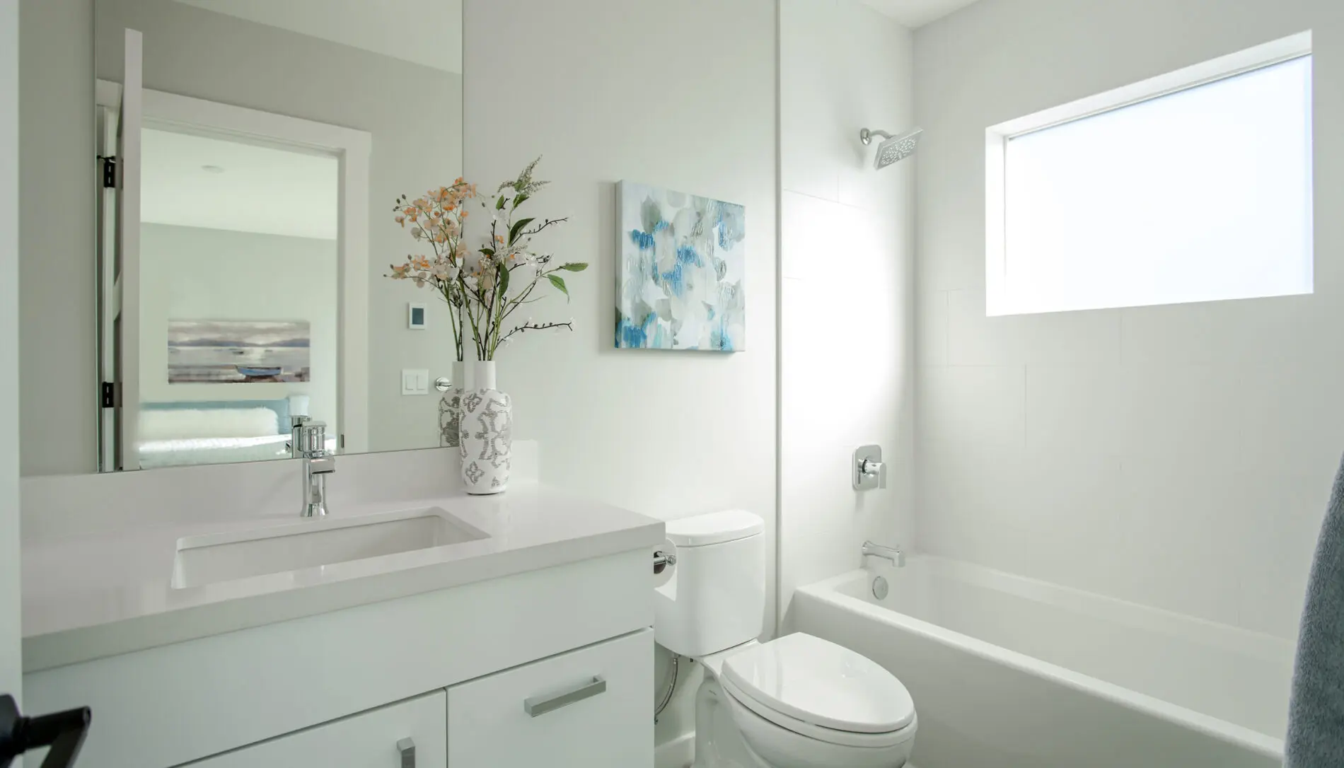 Modern white bathroom in a West Seattle custom home design, featuring a sleek vanity, sink, toilet, bathtub, stylish wall art, vase with flowers, and a frosted window that fills the space with natural light.