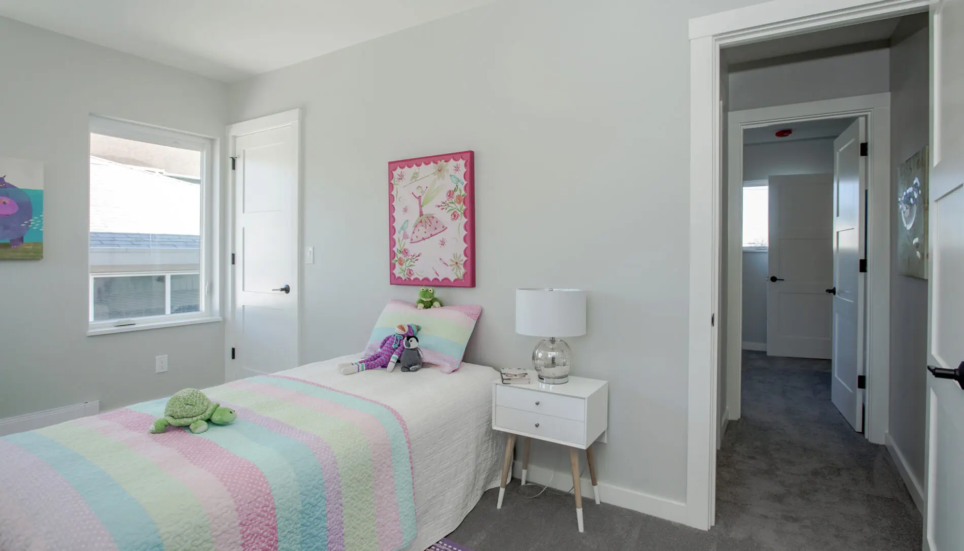 A cozy small bedroom in a West Seattle custom home design features a single bed with a pastel striped quilt, a bedside table and lamp, plush toys on the bed, all set beside a window and an open door.