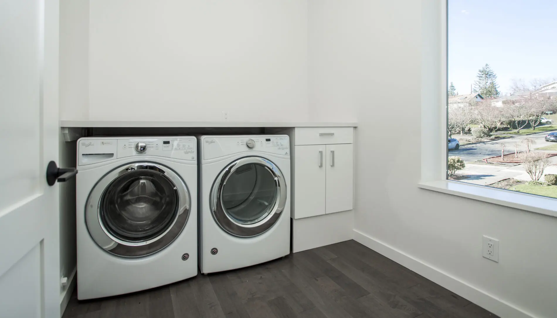 A laundry room with a front-loading washer and dryer next to a white cabinet, large window, and dark wood floor showcases thoughtful West Seattle custom home design.
