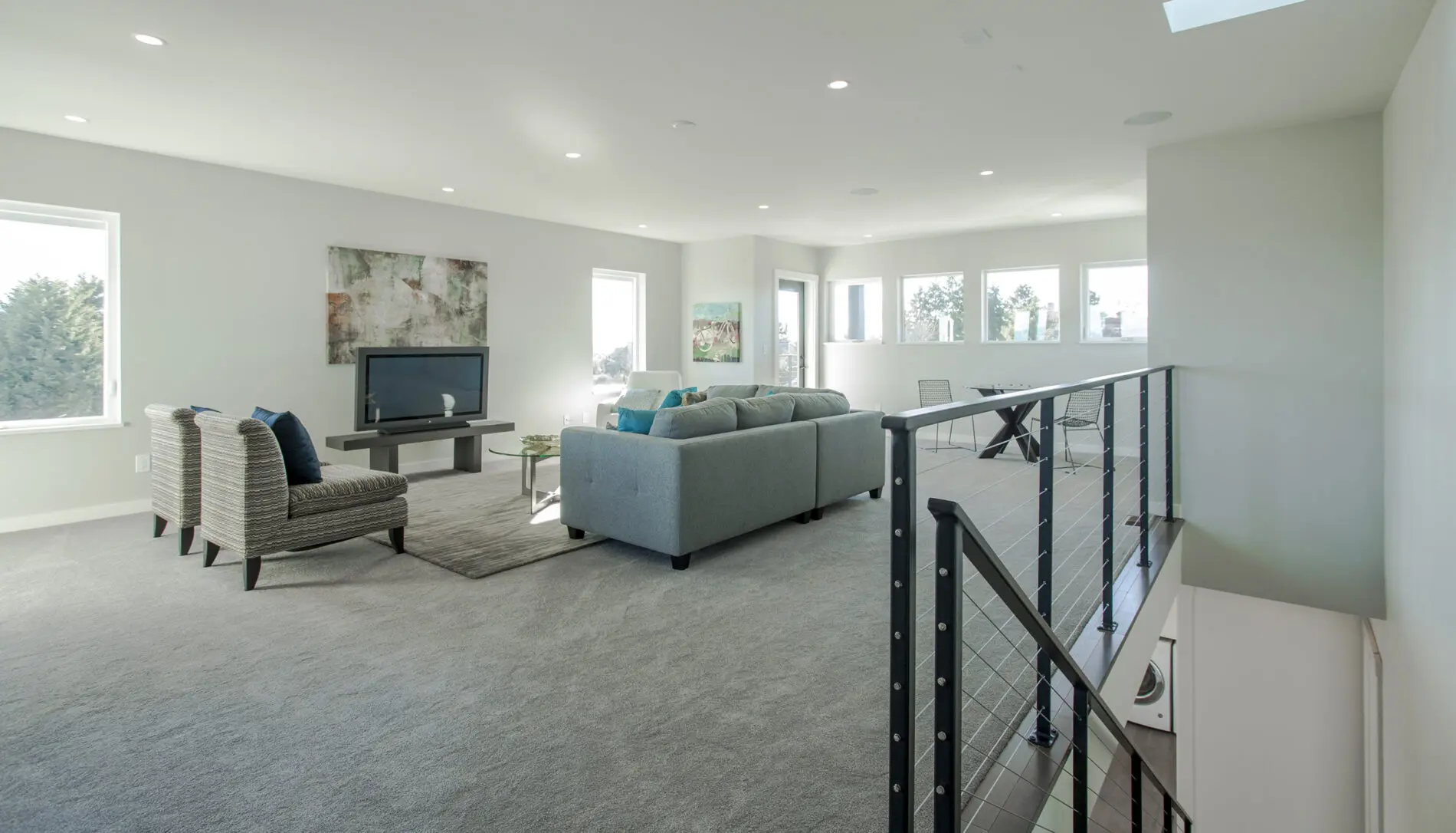 Modern, spacious living room in a West Seattle custom home design, featuring grey carpet, a blue sectional sofa, two armchairs, a TV, artwork on the wall, and large windows providing natural light.