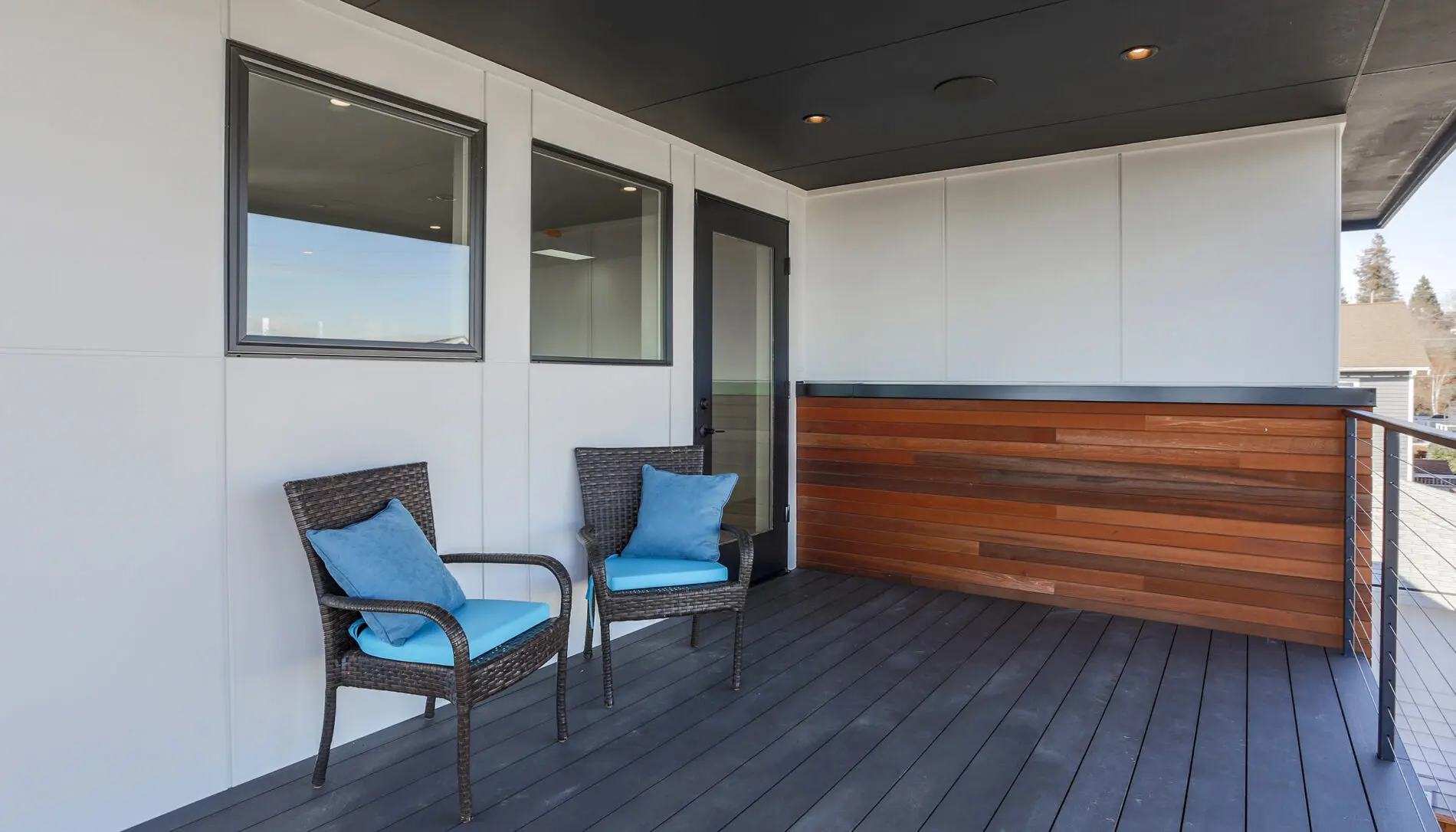 A modern porch with two wicker chairs featuring blue cushions, dark wood flooring, a wooden accent wall, and white paneled walls showcases the elegance of West Seattle custom home design.