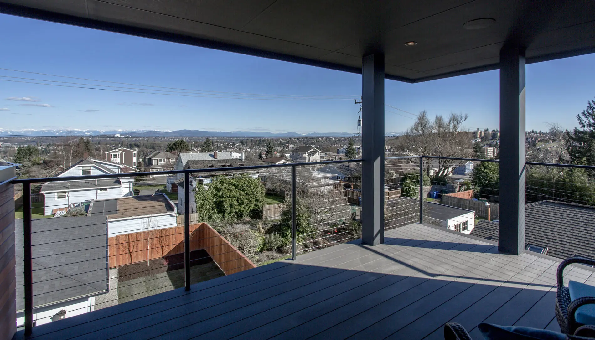 Covered balcony with cable railing in this West Seattle custom home design overlooks suburban rooftops, trees, and a distant city skyline under a clear blue sky.