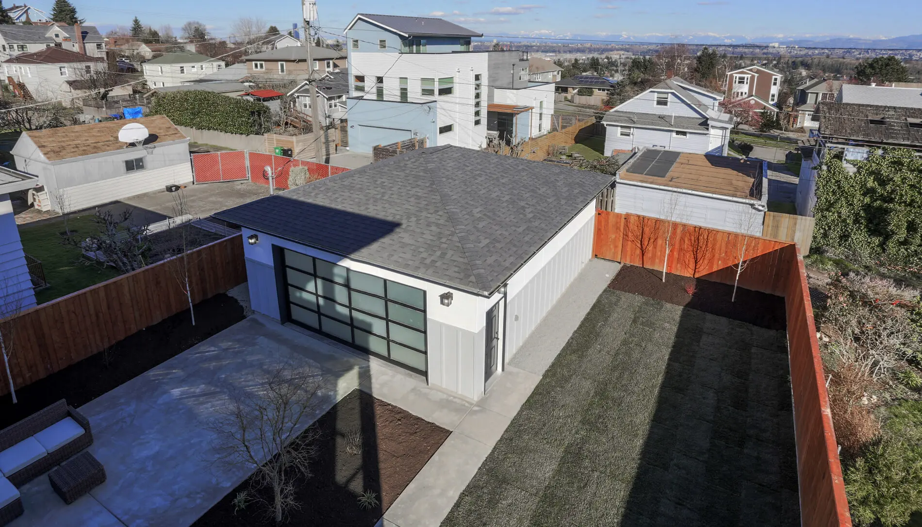 Aerial view of a modern detached garage with a glass-paneled door, set in the fenced backyard of a West Seattle custom home design, featuring a patio and landscaped areas in a residential neighborhood.