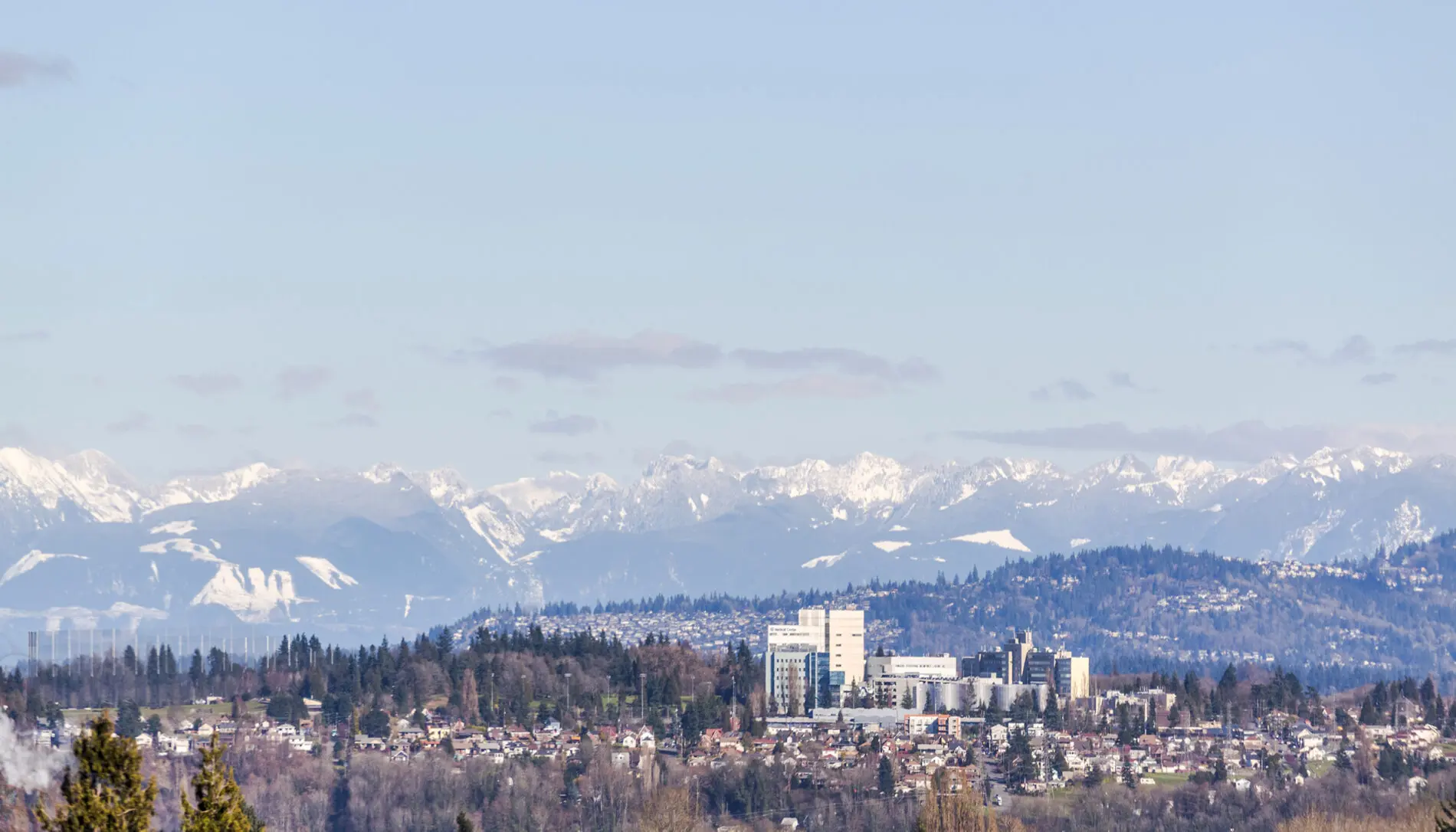 A hospital building is situated on a hill surrounded by residential areas featuring West Seattle custom home design, with snow-capped mountains visible in the background under a clear sky.