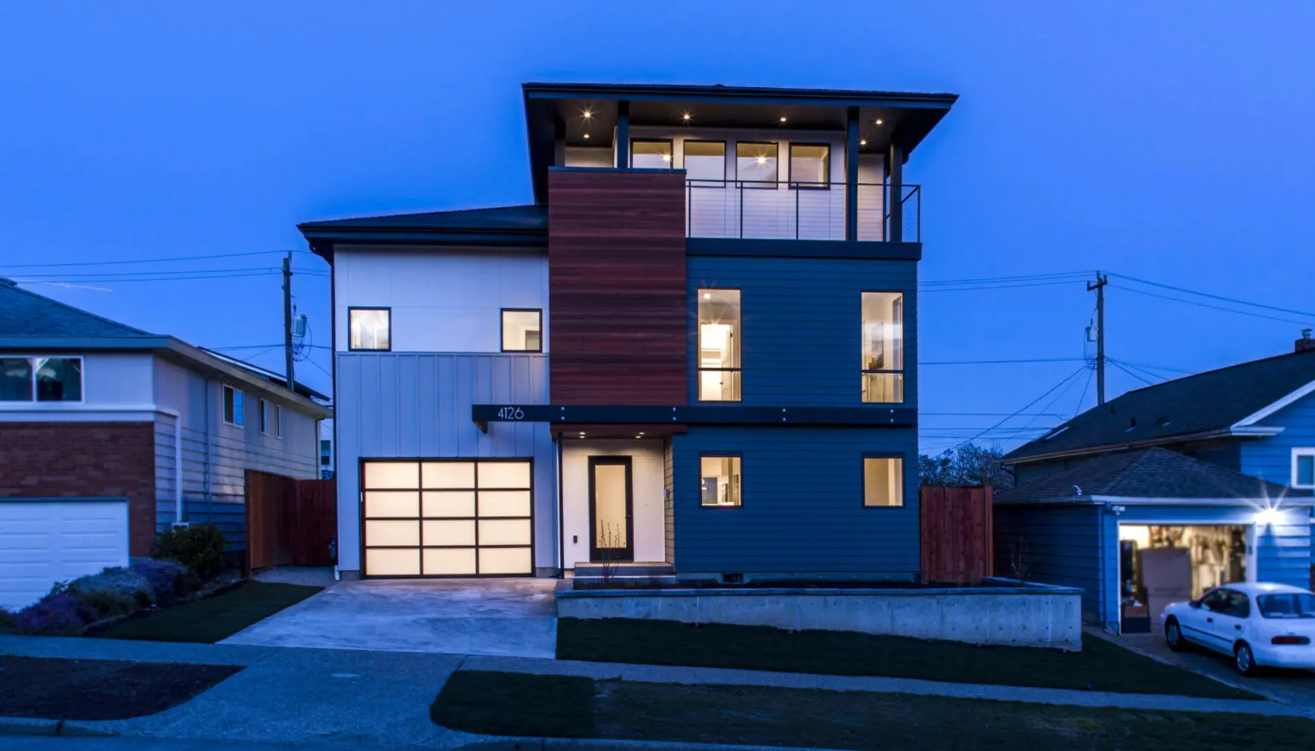 A modern three-story West Seattle custom home design featuring large windows, a wood and blue exterior, and a well-lit garage, photographed at dusk.
