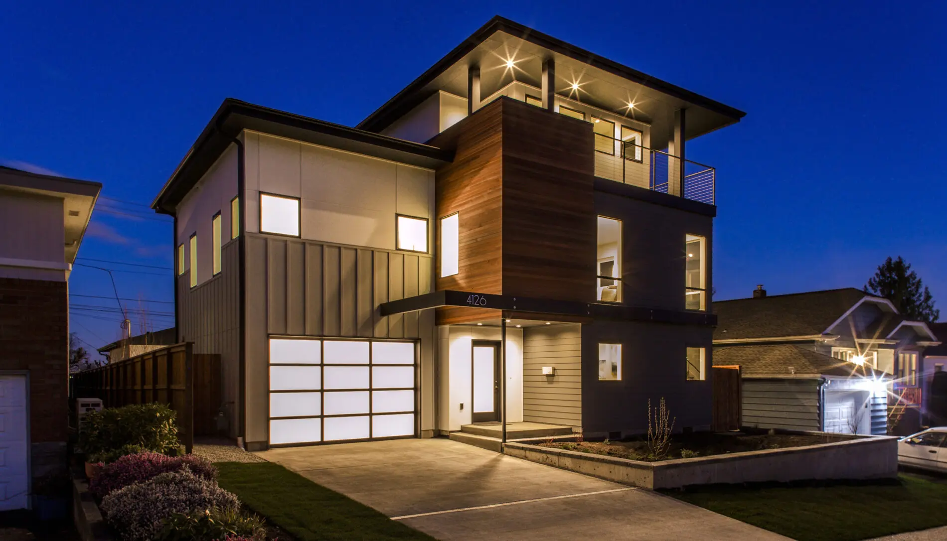 Modern three-story house with large windows and a mix of wood and metal siding, illuminated at night with exterior lighting—a stunning example of West Seattle custom home design.