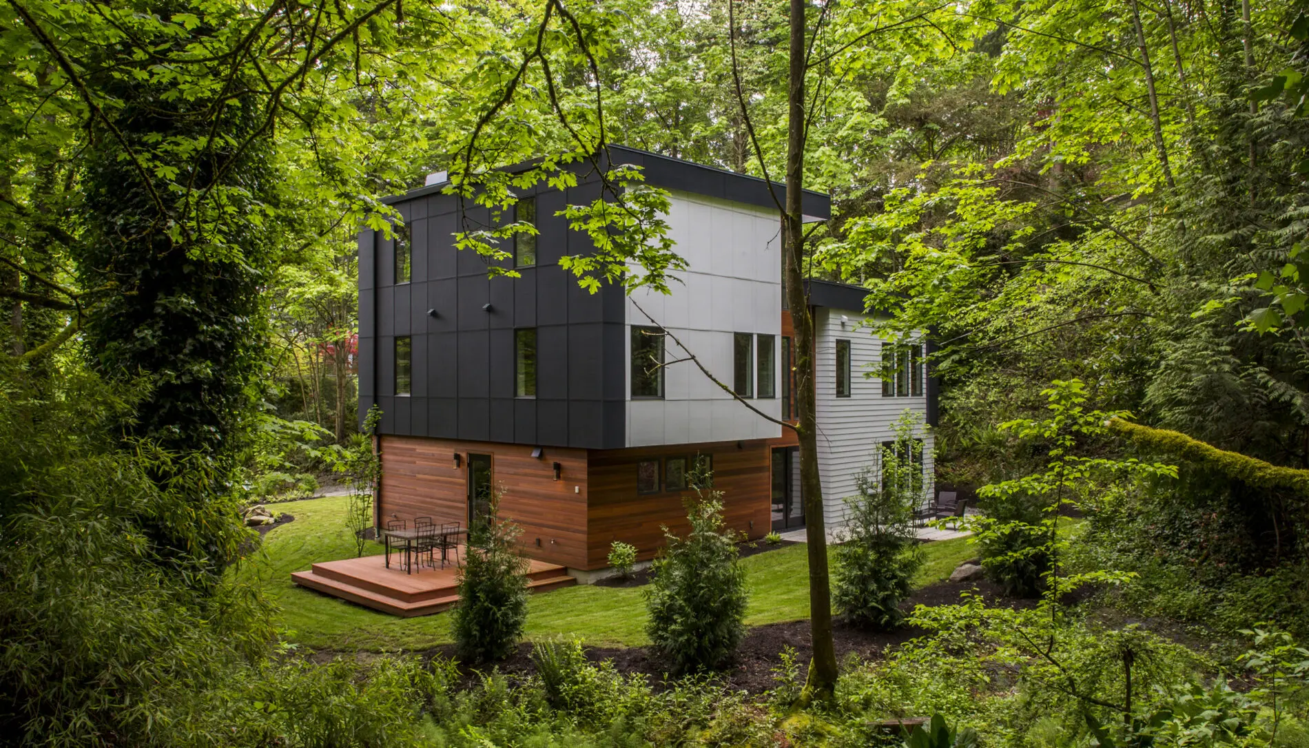 A modern, two-story Mercer Island custom home design with wood and gray panel exterior sits surrounded by dense, green forest and a well-maintained lawn.