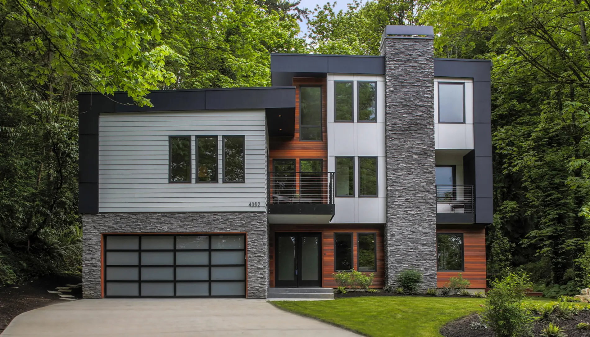 Modern three-story house with large windows, stone and wood accents, a balcony, and a double garage, surrounded by green trees and landscaping—showcasing exceptional Mercer Island custom home design.
