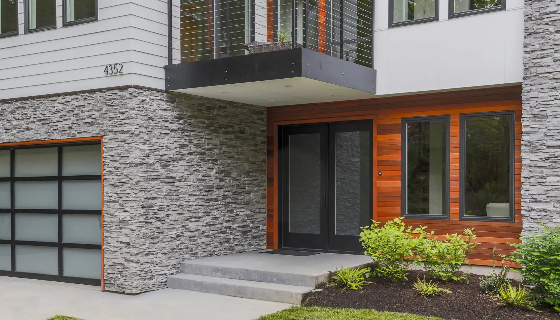 Modern Mercer Island custom home design featuring a glass front door, stone and wood siding, small steps, and a landscaped area with shrubs and mulch.