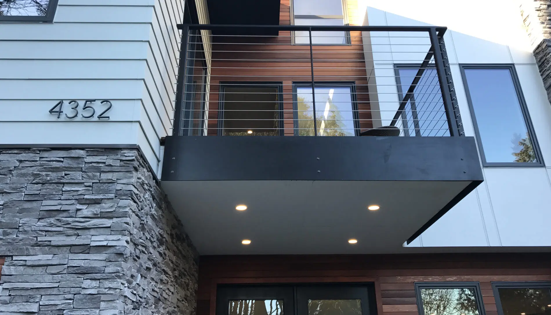 Modern Mercer Island custom home design featuring a mix of stone, wood, and metal finishes, balcony with cable railing, large windows, and address number 4352 mounted on the wall.
