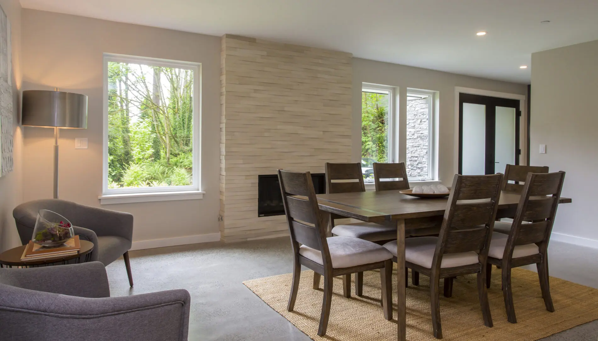 A modern dining room in a Mercer Island custom home design, featuring a dark wooden table, eight chairs, a light-colored rug, gray armchairs, and large windows overlooking greenery.