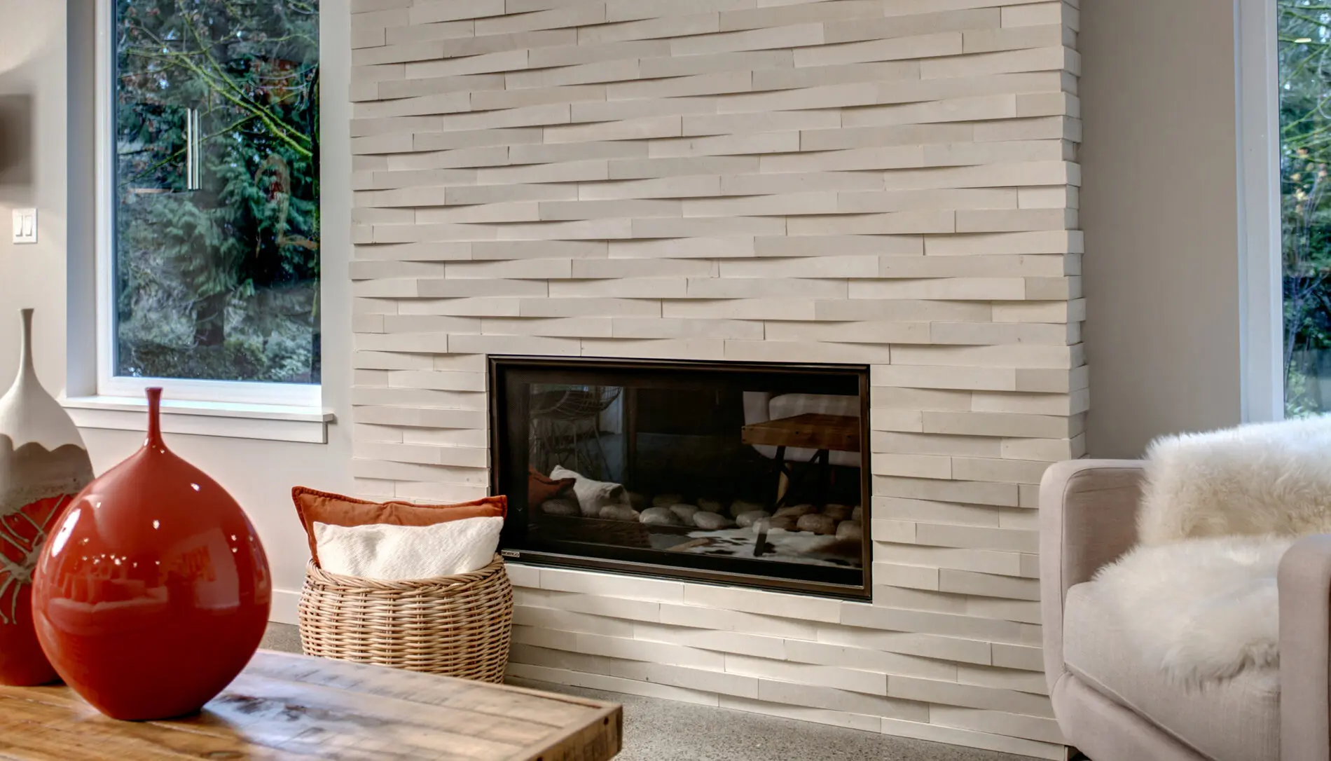 Modern living room with a textured tile fireplace, a woven basket with a pillow, red vase on a wooden table, and beige armchair with white throw—showcasing the elegance of Mercer Island custom home design.
