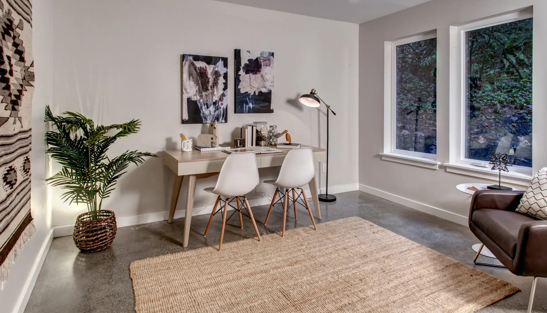 Minimalist home office with a wooden desk, two white chairs, abstract art, a floor lamp, potted plant, woven rug, armchair, and large windows overlooking trees—a perfect example of Mercer Island custom home design.