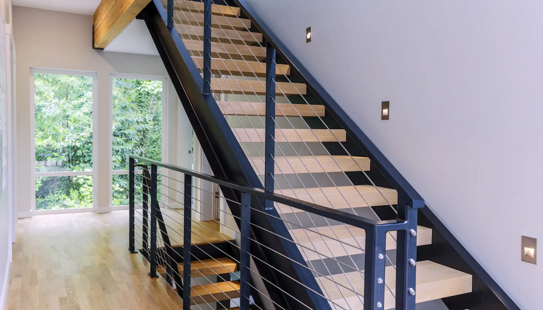 Modern indoor staircase with metal railings, wooden steps, and cable balusters, featured in a stunning Mercer Island custom home design next to large windows with a view of greenery outside.