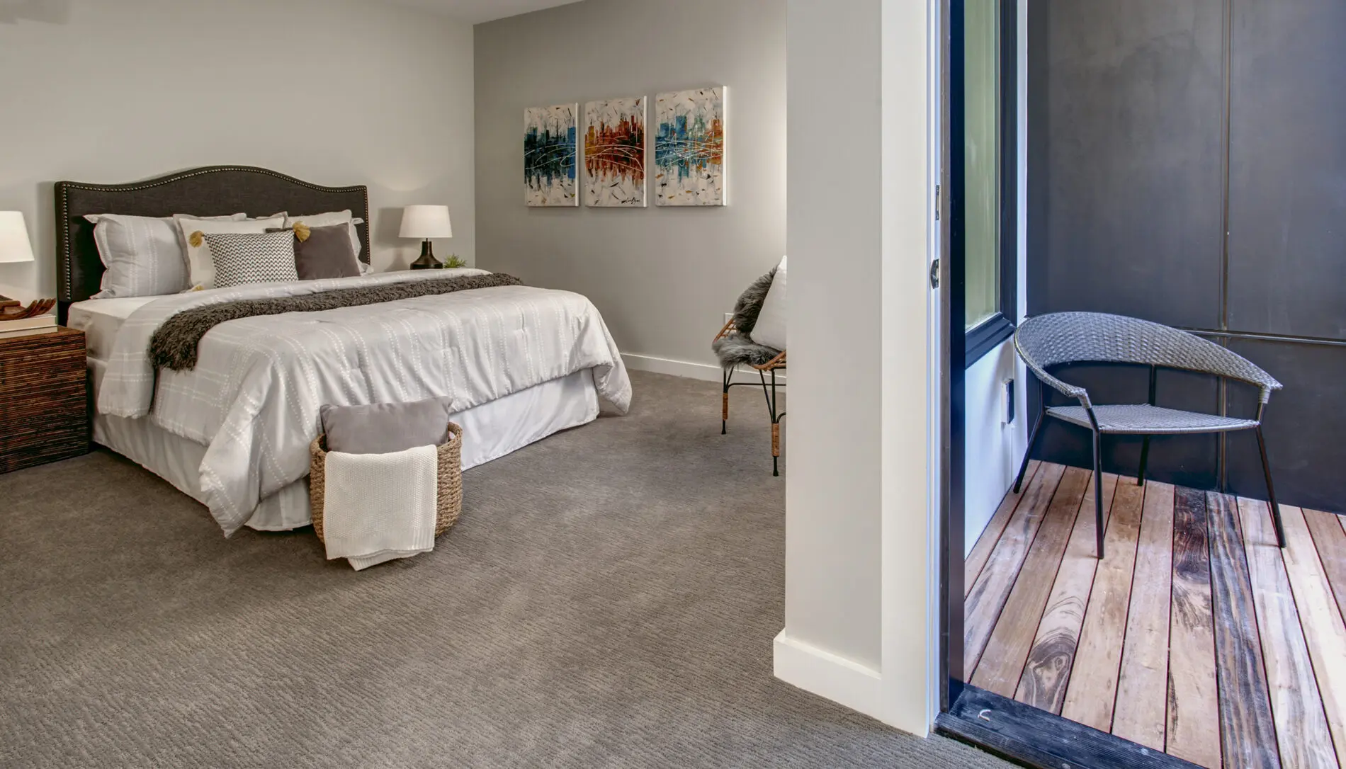 A modern bedroom featuring Mercer Island custom home design, with a large bed, neutral bedding, side tables with lamps, wall art, and access to a small patio with a wicker chair and wooden floor.