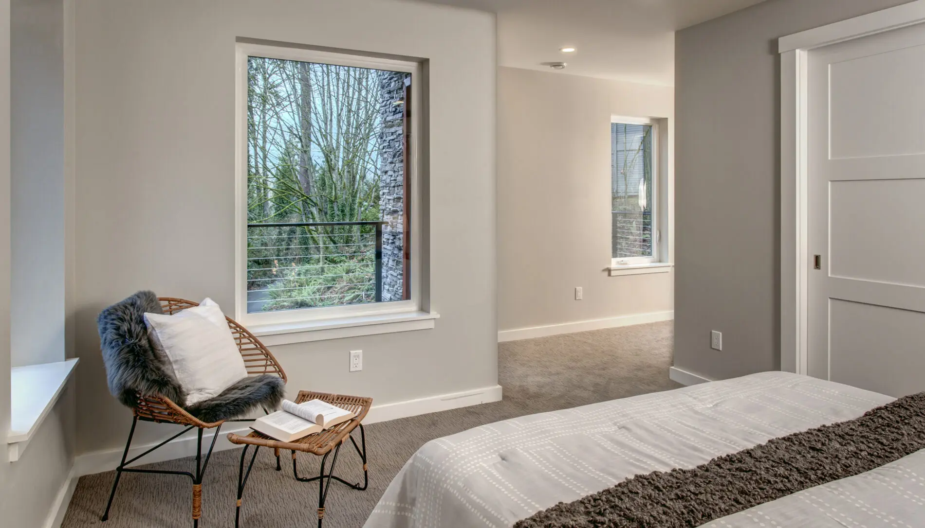 A modern bedroom with neutral walls, a gray blanket on the bed, a wicker chair with cushion and fur throw, a small table with an open book, and large windows revealing trees—reflecting Mercer Island custom home design.