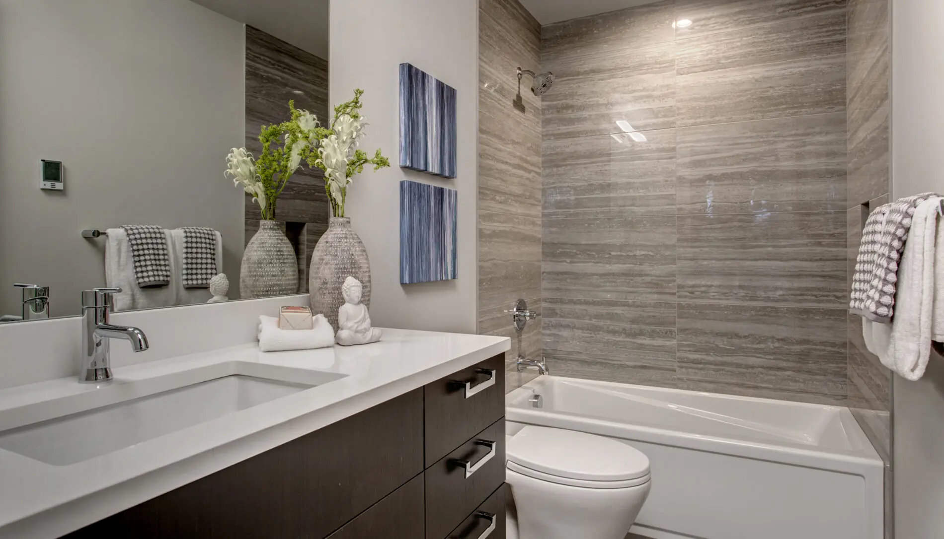 Modern bathroom with a white sink, dark vanity, bathtub with gray tile walls, toilet, towels, vases of flowers, and two blue wall art pieces—showcasing elegant Mercer Island custom home design.