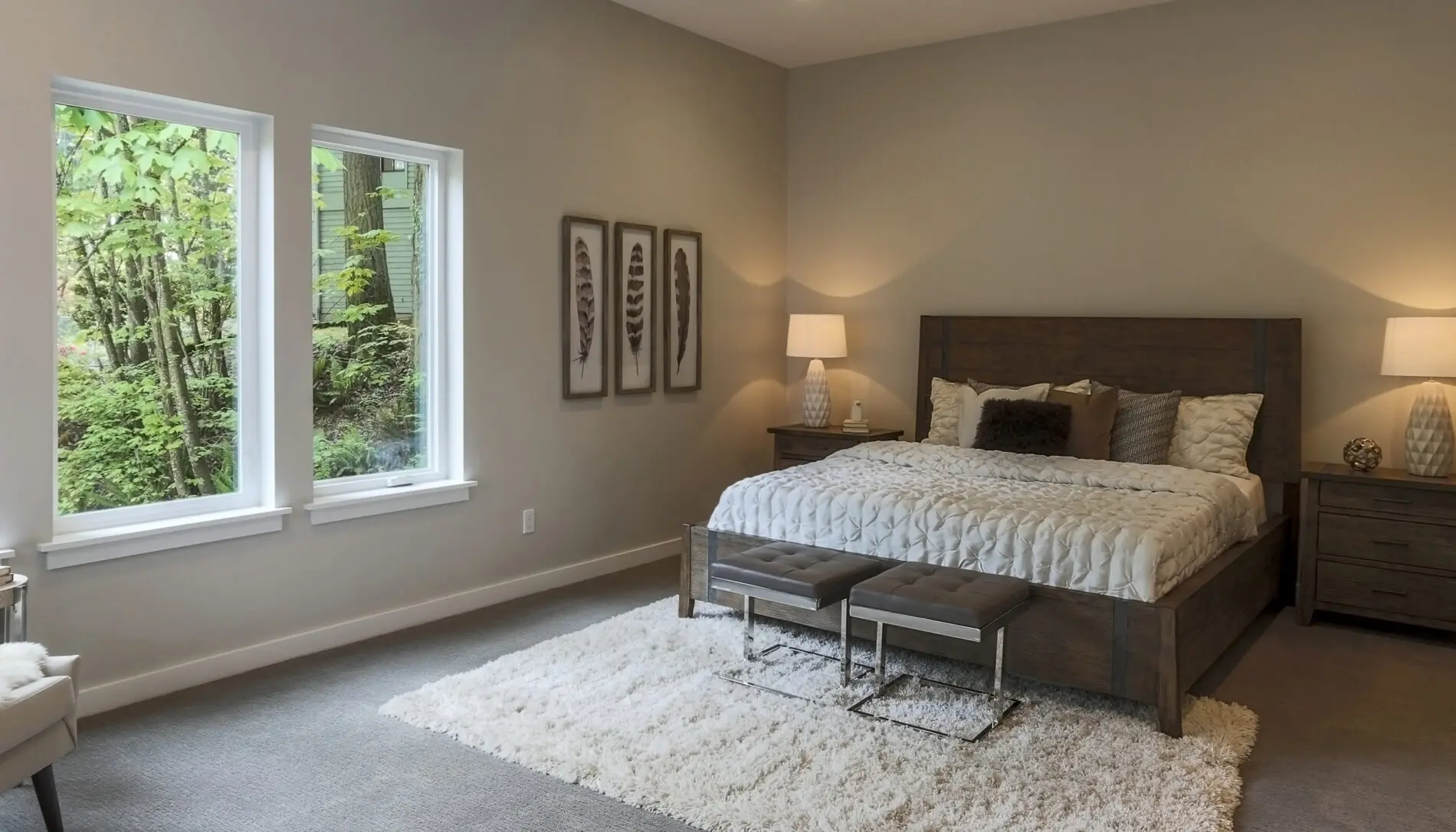 A modern bedroom in a Mercer Island custom home design features a large bed, two benches at the foot, nightstands with lamps, three framed art pieces, and two windows revealing lush green trees outside.