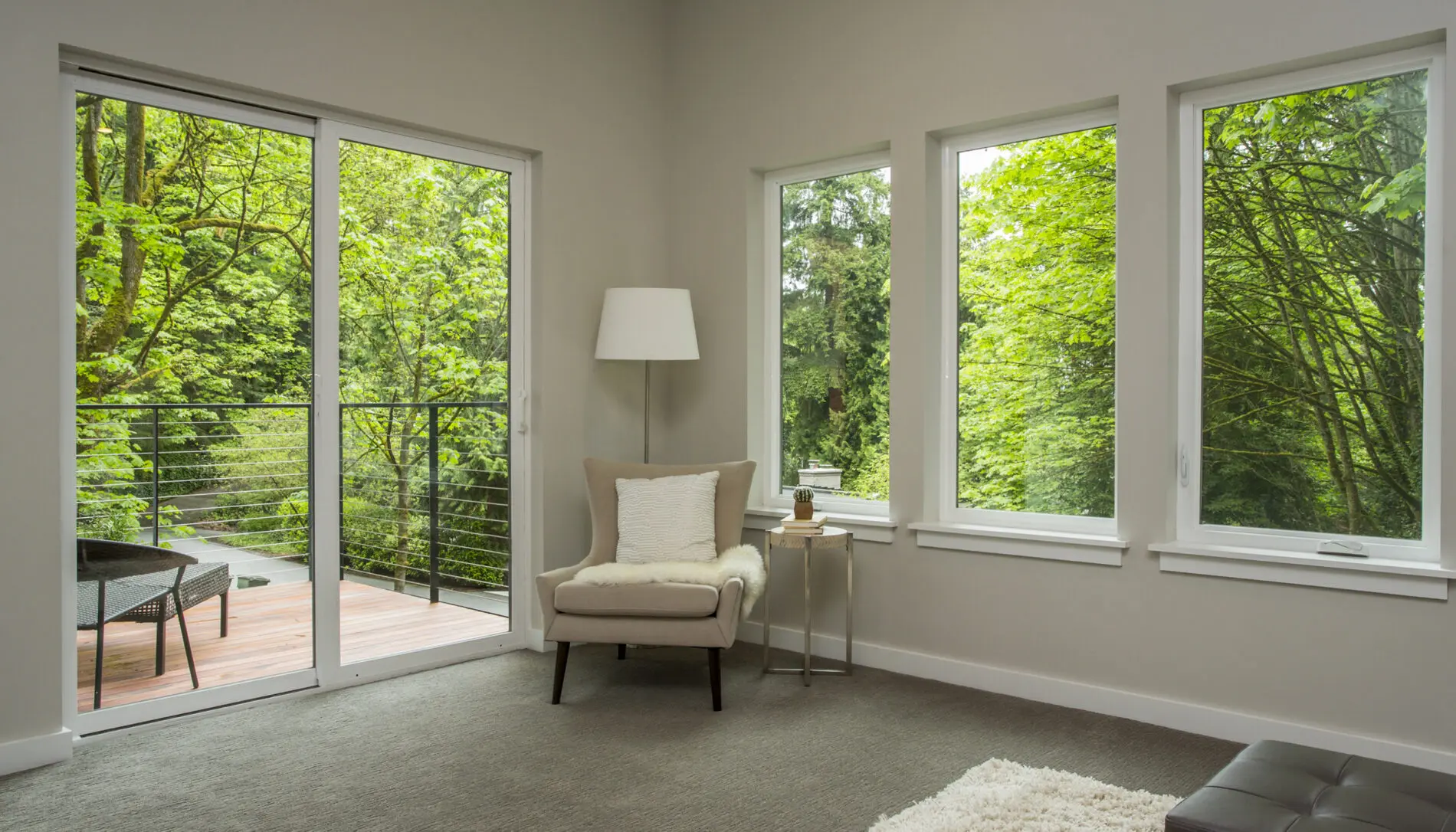 A modern living room with a beige armchair, floor lamp, and side table, showcases large windows and a glass door overlooking a green, wooded deck area—a beautiful example of Mercer Island custom home design.