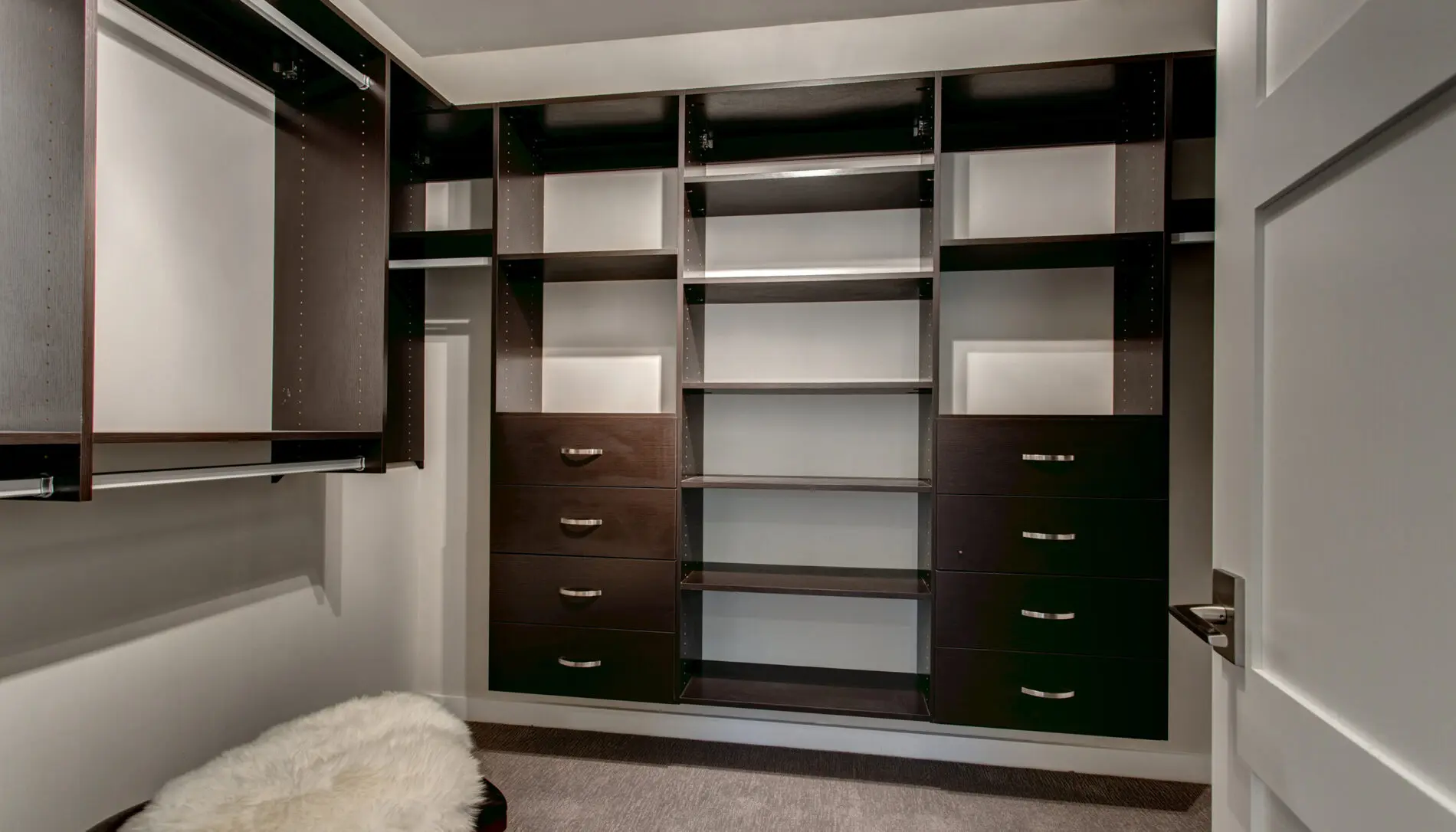 Empty walk-in closet with dark wood shelving, multiple drawers, hanging rods, and a stool with a white fur cover on a carpeted floor—showcasing refined Mercer Island custom home design.