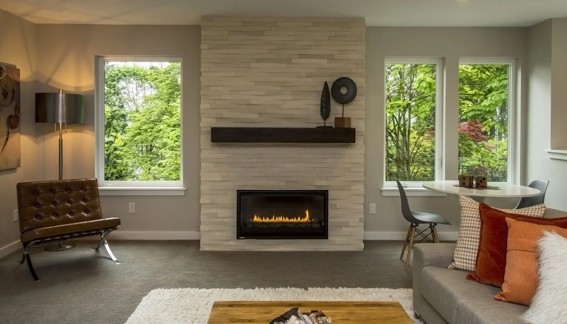 Modern living room with a gas fireplace, neutral decor, large windows showing trees, a brown chair, gray sofa, and cozy dining area—showcasing Mercer Island custom home design at its finest.