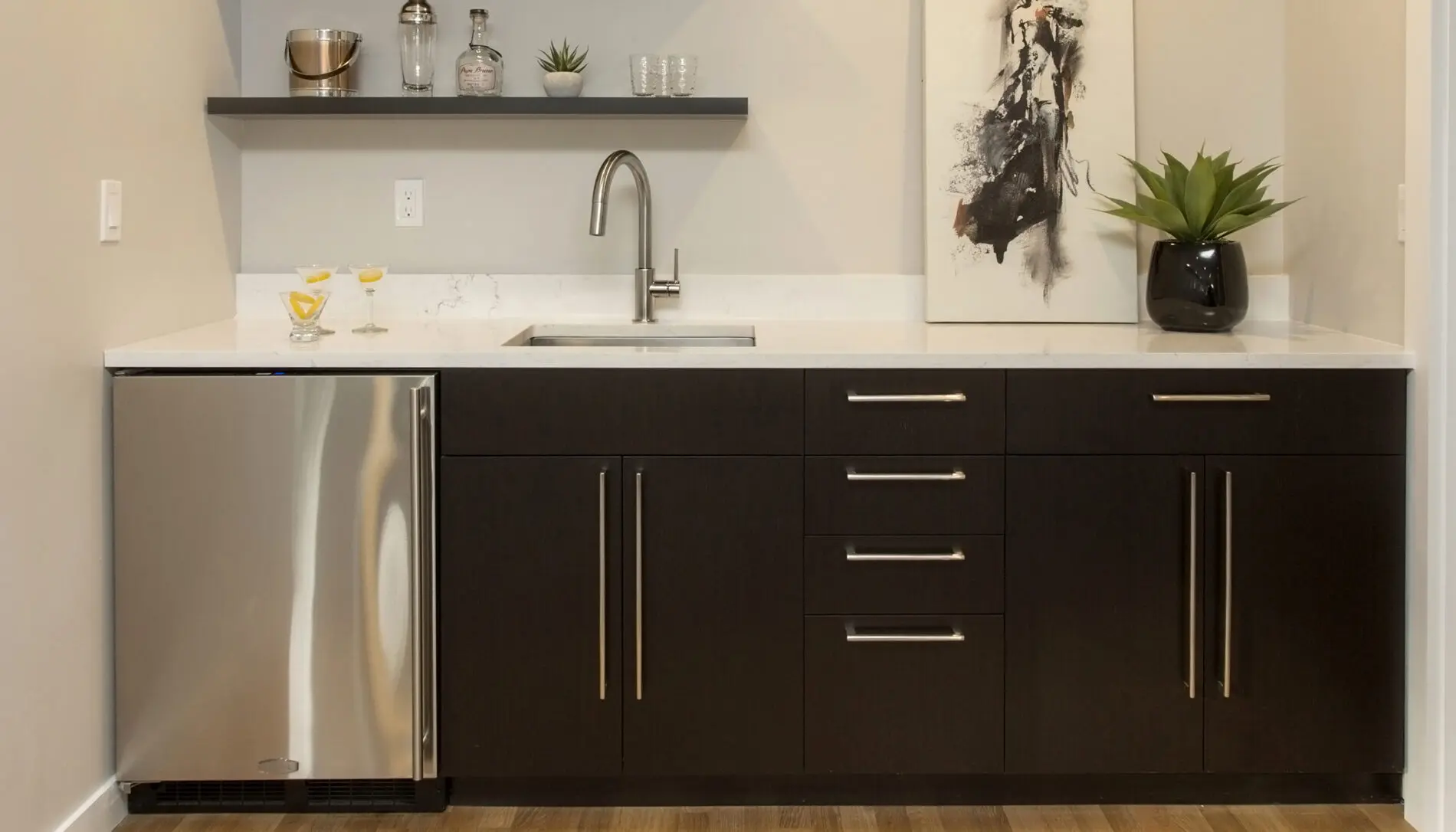 Modern kitchenette with dark cabinets, a stainless steel mini fridge, a sink, open shelving with decor, and a plant on the counter—showcasing Mercer Island custom home design flair. Abstract art hangs above the counter.