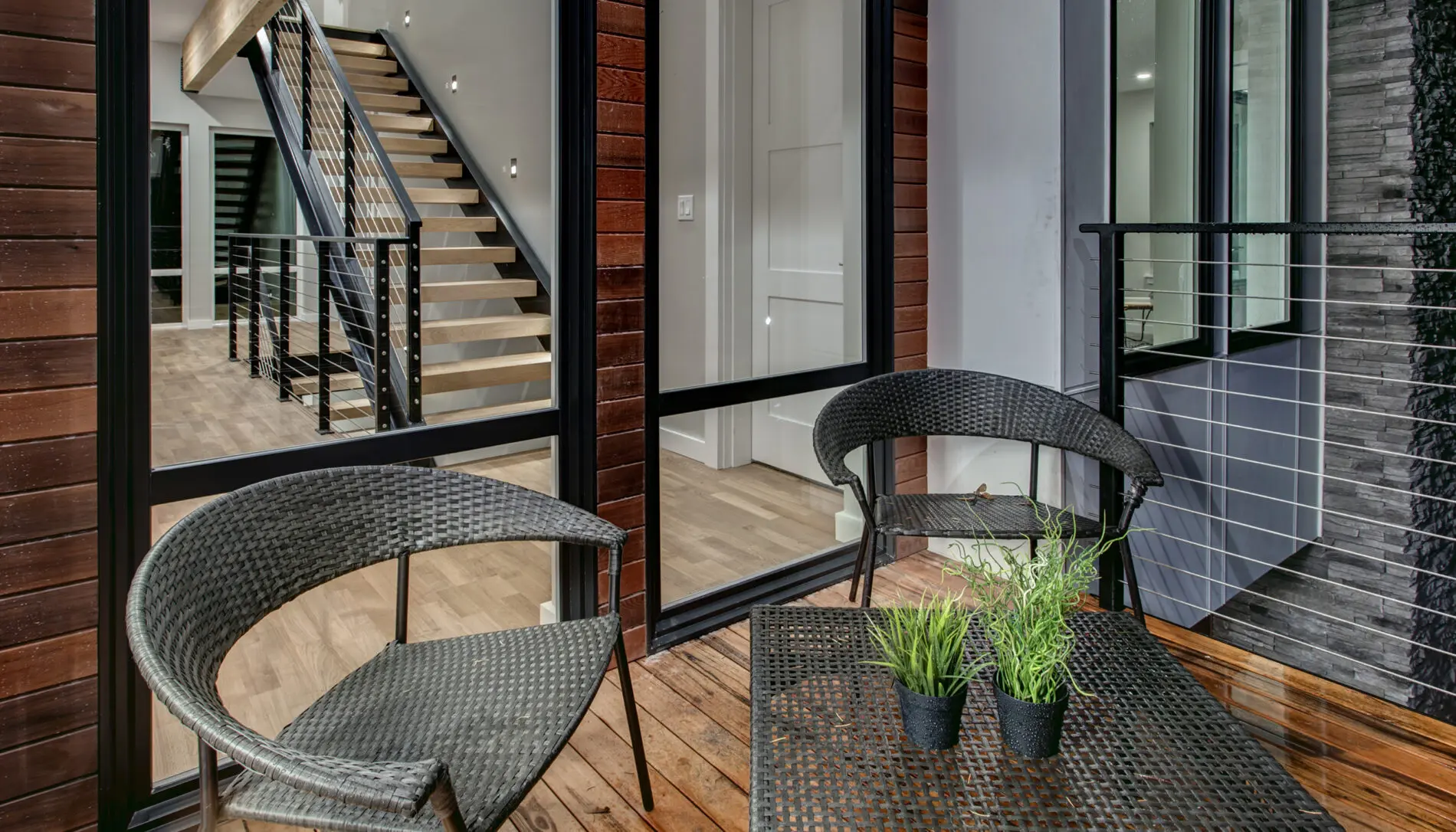 Modern outdoor patio with two wicker chairs, a glass table with potted plants, and a view of an indoor staircase through large windows—showcasing elegant Mercer Island custom home design.