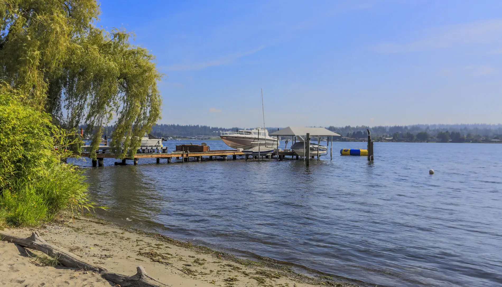 A small dock with a boat and covered lift extends into the water from a sandy shore with trees, greenery, and Mercer Island custom home design touches under a clear blue sky.