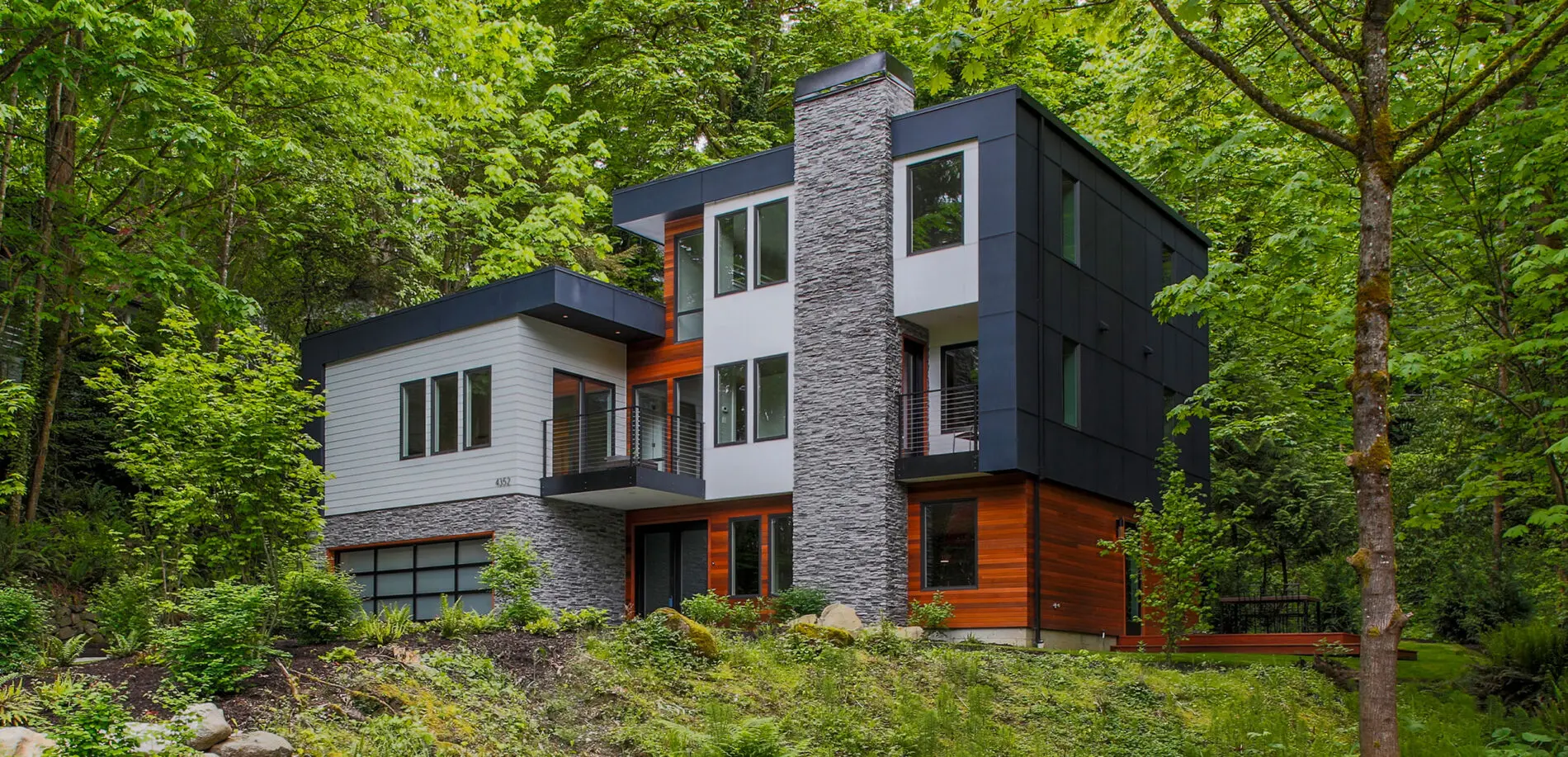 Modern new construction two-story house with stone, wood, and black panel exterior, surrounded by dense green trees and vegetation.