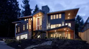 Modern two-story house exemplifying Pacific Northwest luxury residential architecture, featuring large windows, a wood and stone exterior, sloped roof, outdoor stairs, and illuminated interior lighting at dusk.