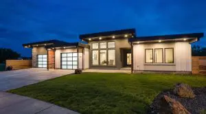 Modern single-story house at dusk features Pacific Northwest luxury residential architecture with large windows, illuminated exterior lights, flat roofs, and a landscaped front yard with concrete driveway and wood accents.