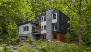 Modern two-story house showcasing Pacific Northwest luxury residential architecture, with stone, wood, and black panel exterior, large windows, and balconies nestled among dense green trees and vegetation.
