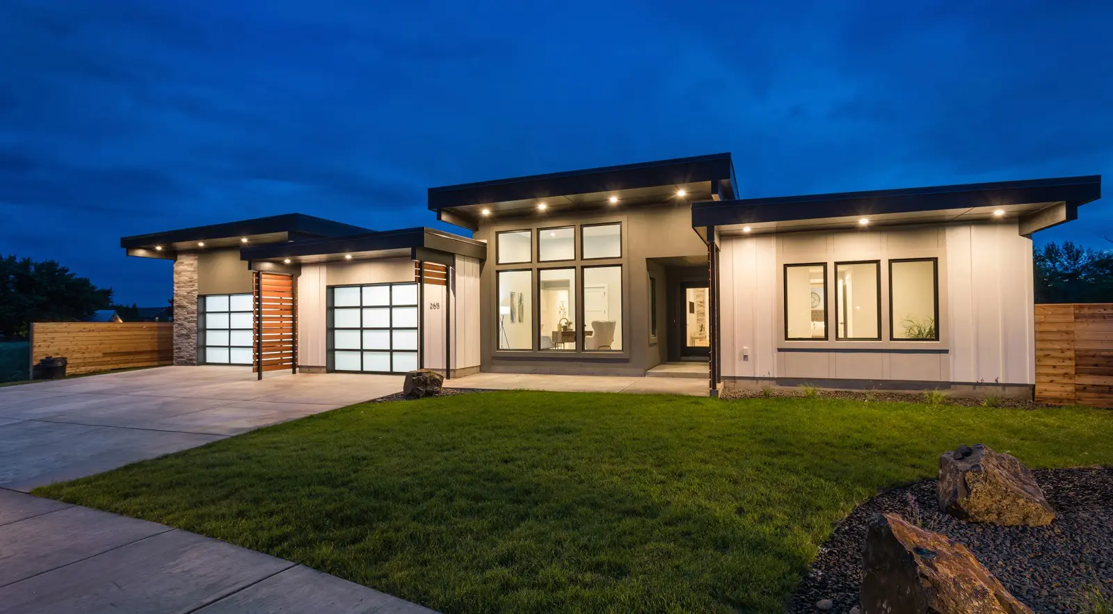 A modern single-story house featuring Walla Walla custom home design, with large windows, flat roofs, illuminated exterior lights, a two-car garage, and a neatly manicured lawn at dusk.