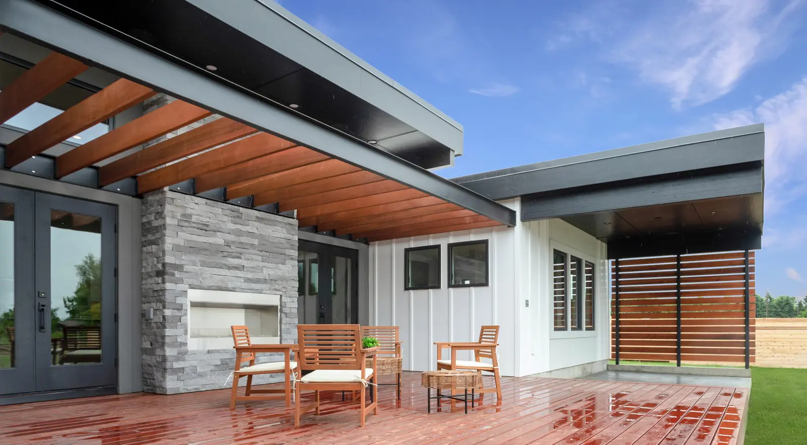 Modern house exterior with a wooden patio, outdoor seating, and a stone fireplace showcases Walla Walla custom home design, featuring horizontal wood slats beneath a partly cloudy sky.