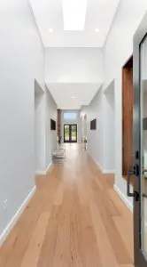 A modern hallway with light wood flooring, white walls, recessed lighting, and a glass door showcases signature Walla Walla custom home design, leading to a living area with large windows and stunning outdoor views.