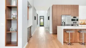 Modern kitchen with wood cabinetry, stainless steel appliances, white countertops, two barstools, and light wood flooring—showcasing Walla Walla custom home design—opens to a hallway with built-in shelves and a view of the outdoors.