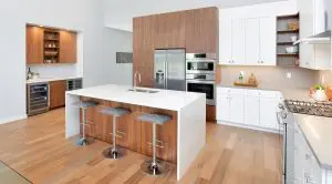 Modern kitchen with white countertops, wood cabinets, stainless steel appliances, a central island with three stools, and a separate bar area on the left—showcasing refined Walla Walla custom home design.
