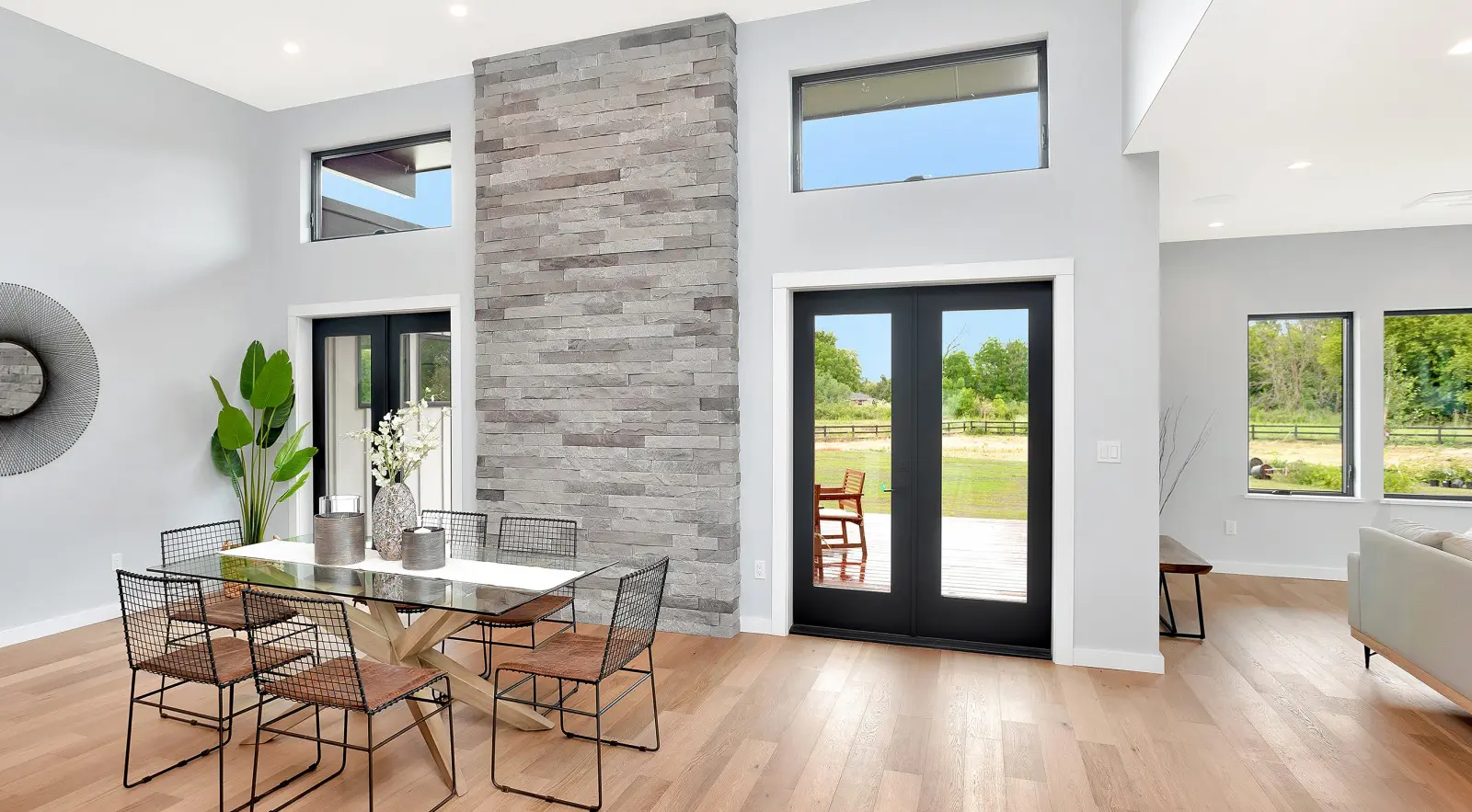 Modern dining area with a stone accent wall, glass table, metal chairs, large windows, and glass double doors overlooking a green outdoor landscape—showcasing the elegance of Walla Walla custom home design.