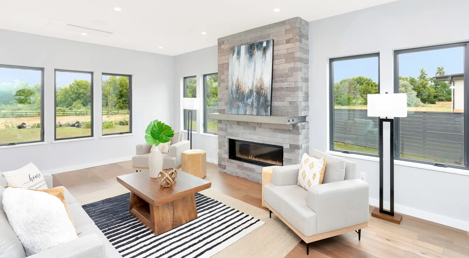 Modern living room with large windows, light-colored furniture, a striped rug, a wooden coffee table, and a stone fireplace topped with abstract art—showcasing the elegance of Walla Walla custom home design.